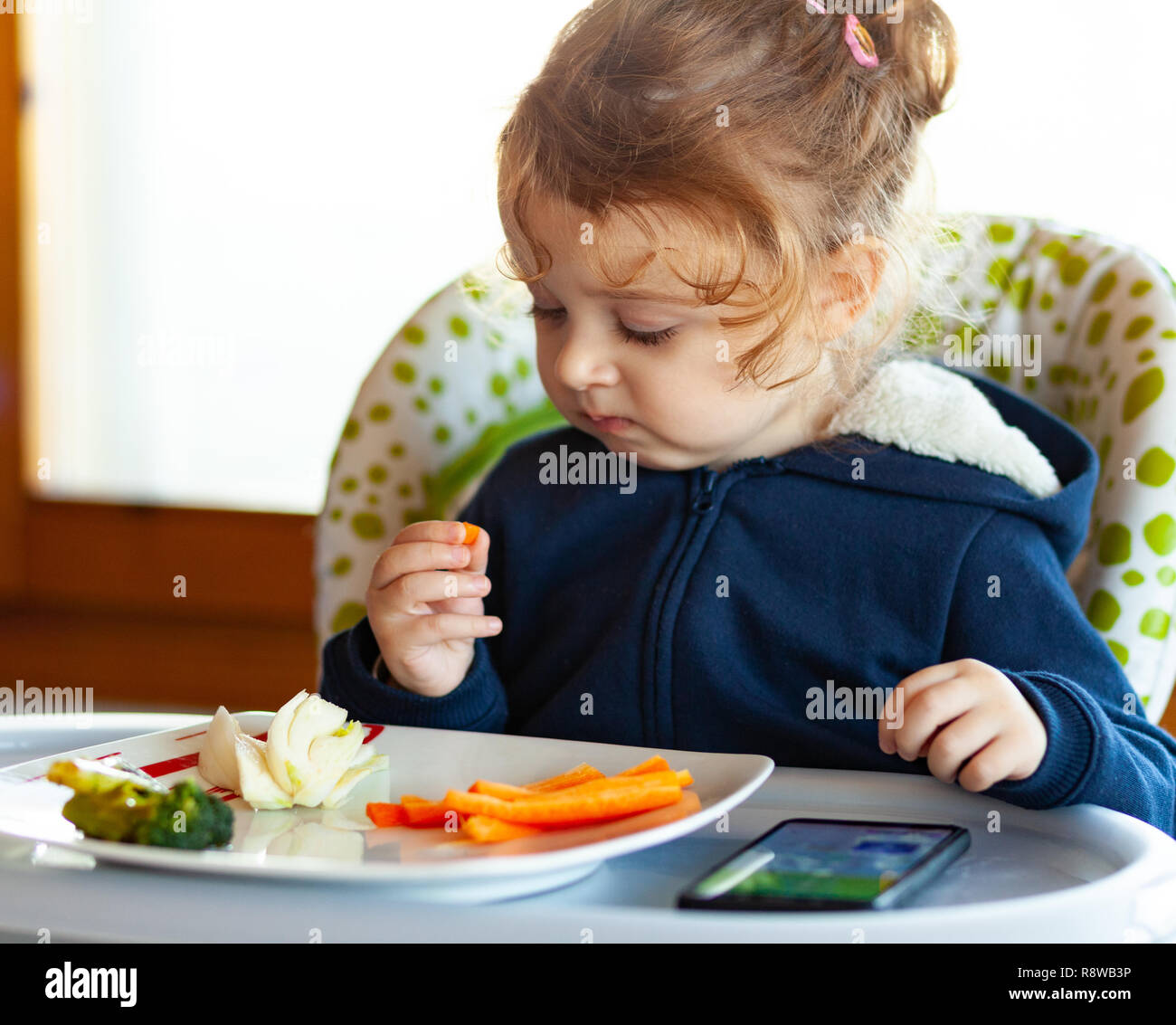 Toddler eats in the high chair while watching movies on the mobile phone. Many children do not want to eat if they do not watch the mobile device or t Stock Photo