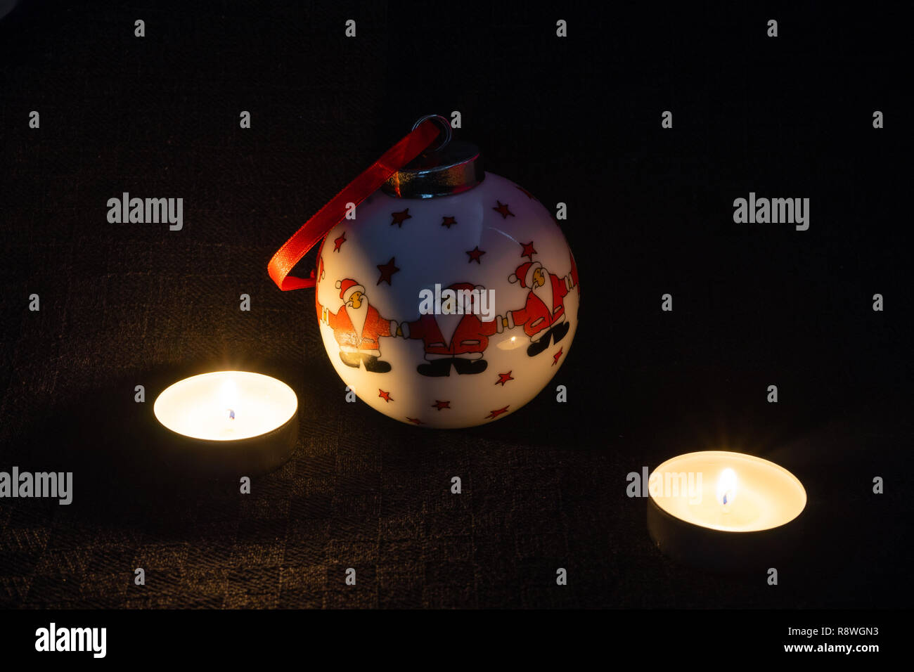 A decorative Christmas bauble depicting three Santa Clauses with two lit candles next to it against black background- Christmas concept Stock Photo
