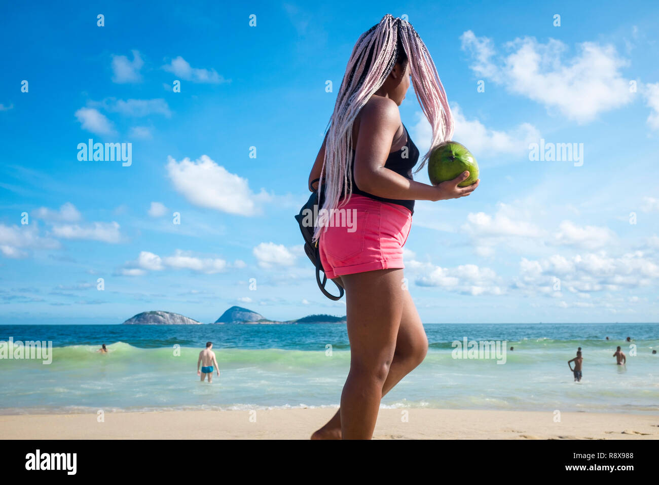 RIO DE JANEIRO - JANUARY, 2018: A young woman with pink braided hair walks with a fresh green coconut on the shore of Ipanema Beach. Stock Photo