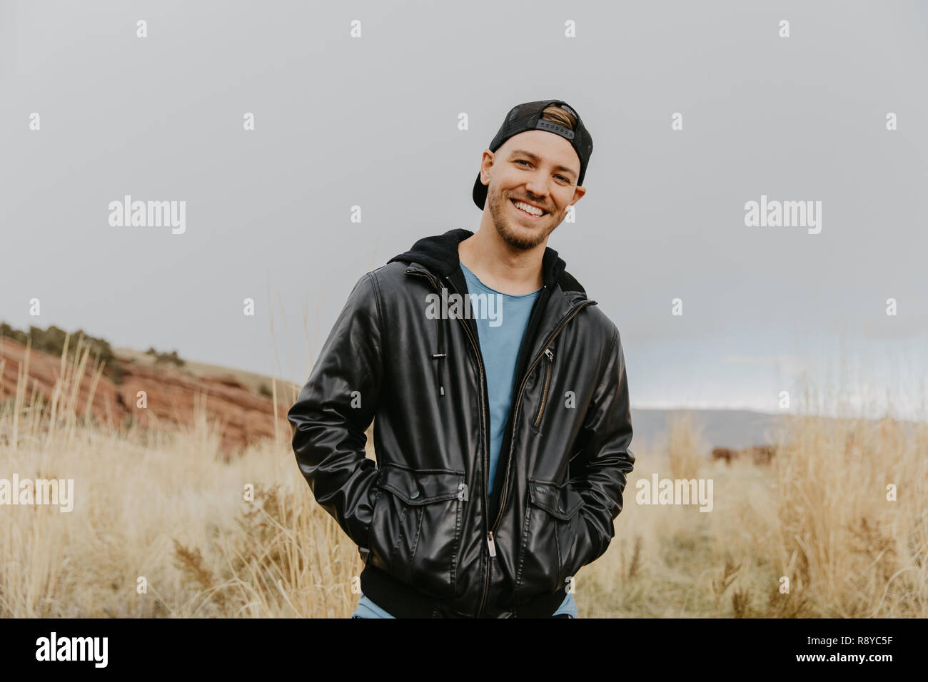 Portrait of Young Good Looking Handsome Man Smiling with Backwards Hat, Leather Jacket Outside in Isolated Rural Field of Tall Grass in the Mountain Stock Photo