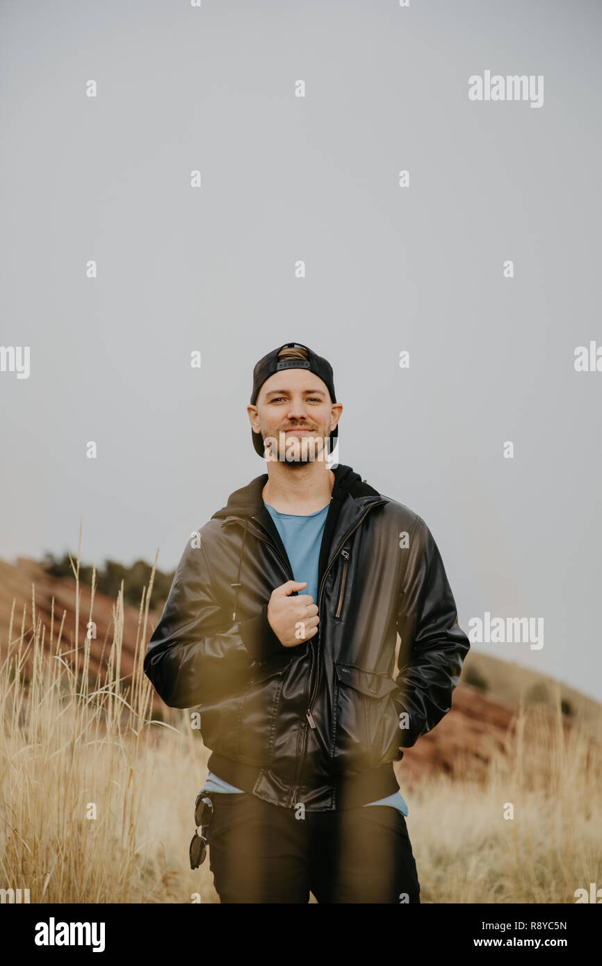 Portrait of Young Good Looking Handsome Man Smiling with Backwards Hat, Leather Jacket Outside in Isolated Rural Field of Tall Grass in the Mountain Stock Photo