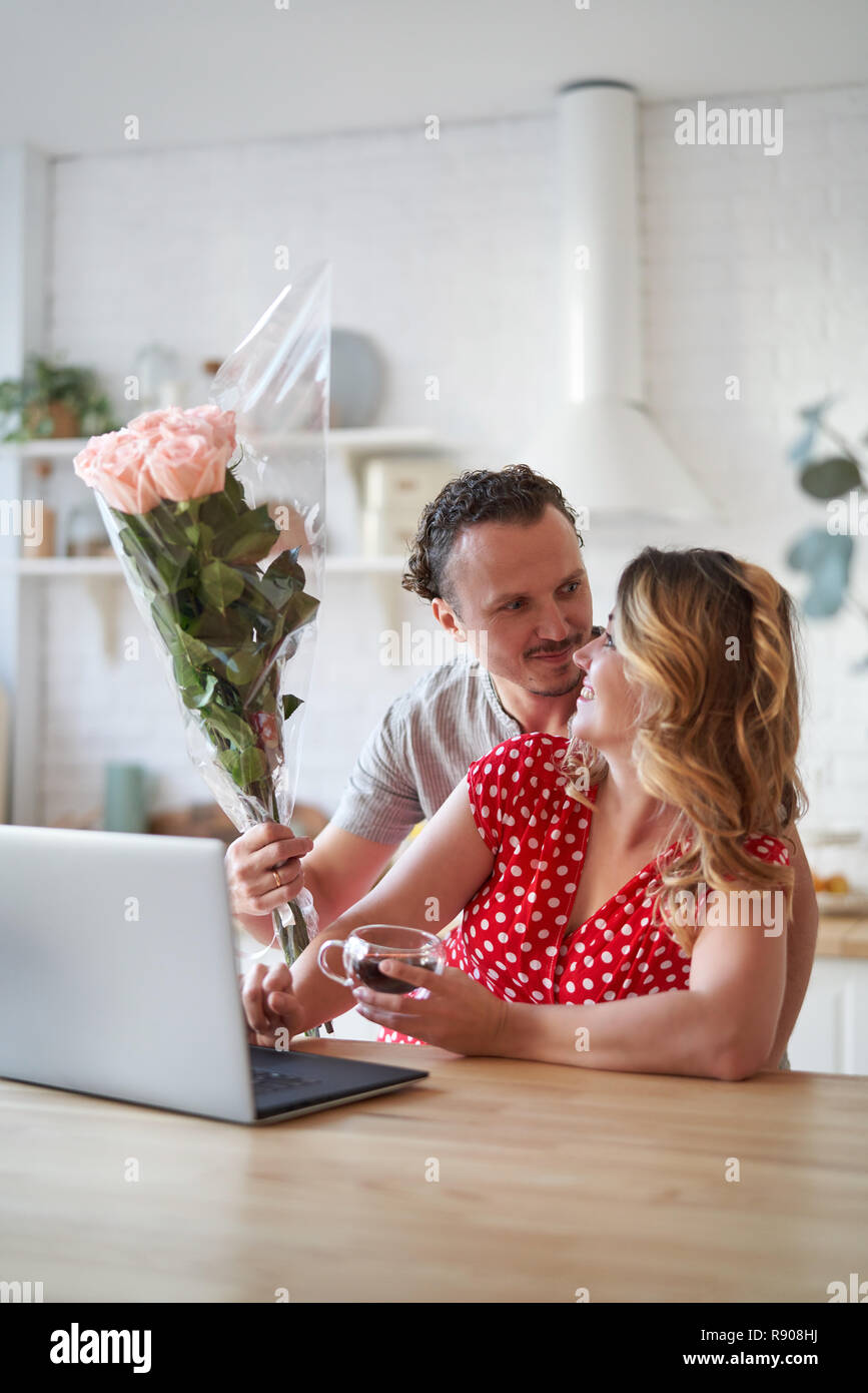Surprise. Beautiful romantic couple in kitchen. Young man is presenting flowers to his beloved. Feel of happiness. Stock Photo