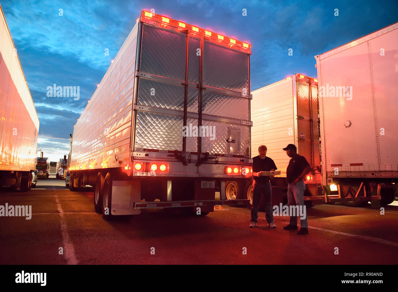 Two truck drives checking dispatch papers while standing next to truck trailers in a large parking lot at night. Stock Photo