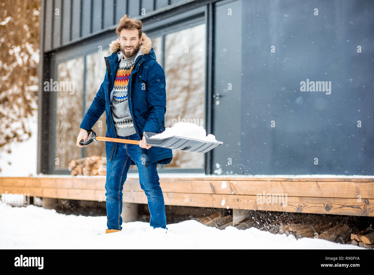 Handsome man in winter clothes cleaning snow with a shovel near the modern house in the mountains Stock Photo