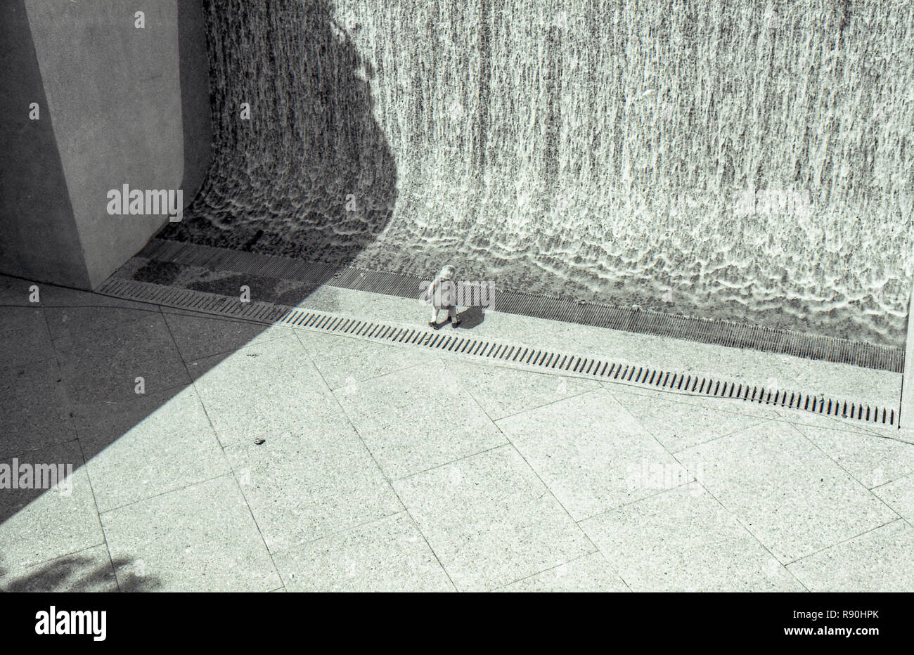 Sydney Australia, March 13th 1977: A young girl plays at the base of the Sydney Square Waterfall designed as part of the greater Square plan by Ridley Smith. In July 1977 the word Eternity was embedded in the pavement at the base of the waterfall to commemorate the life of Arthur Stace. Ending with his death in July 1967 Arthur Stace wrote a single word sermon Eternity in chalk on the pavements of Sydney in beautiful copper place writing for 34 years. Stace was further remembered when the word Eternity was lit up on the Sydney Harbour Bridge as part of the year 2000 New Years Eve celebrations. Stock Photo