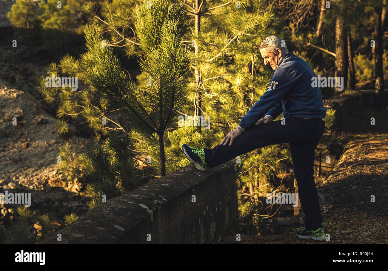 Profile shot of a senior with white hair in sportswear doing stretching exercises in a park at sunset Stock Photo