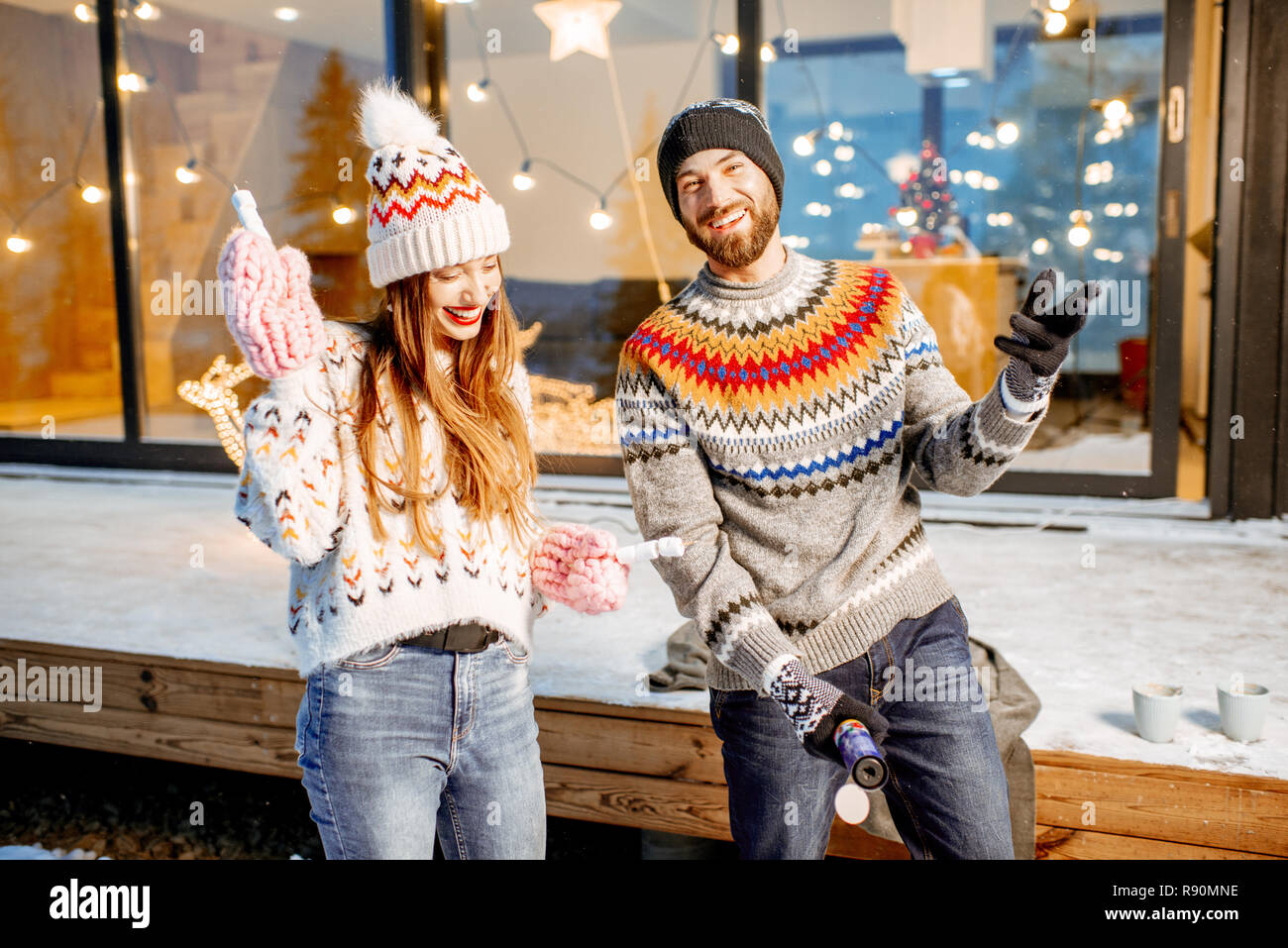 Young happy couple dressed in sweaters celebrating winter holidays in front of a beautiful decorated house in the mountains Stock Photo