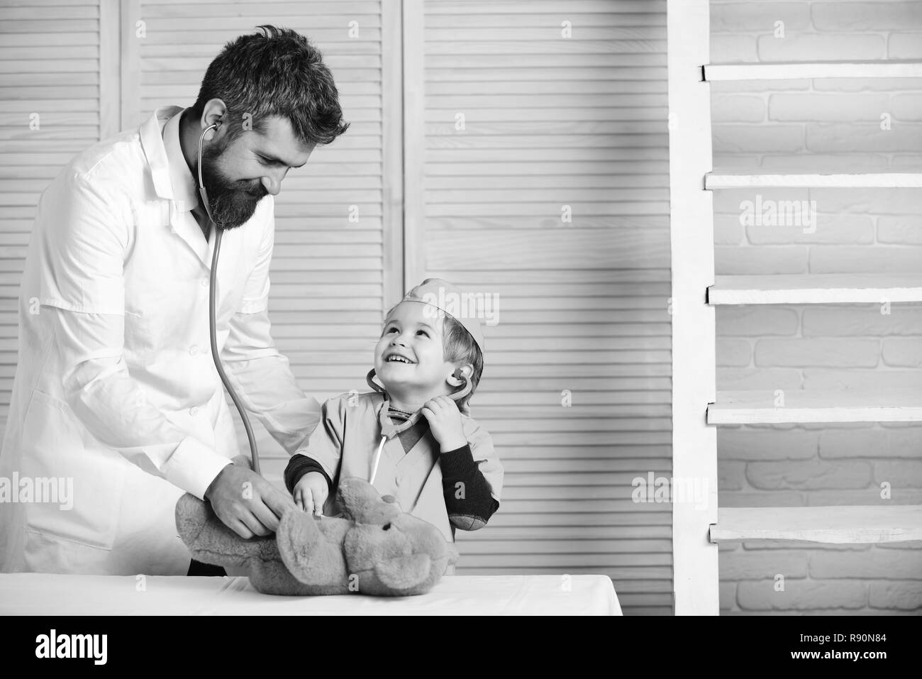 Man with beard and boy hold stethoscope on wooden background. Healthcare and childhood concept. Vet and little assistant examine teddy bear. Father and kid with happy faces play doctor Stock Photo