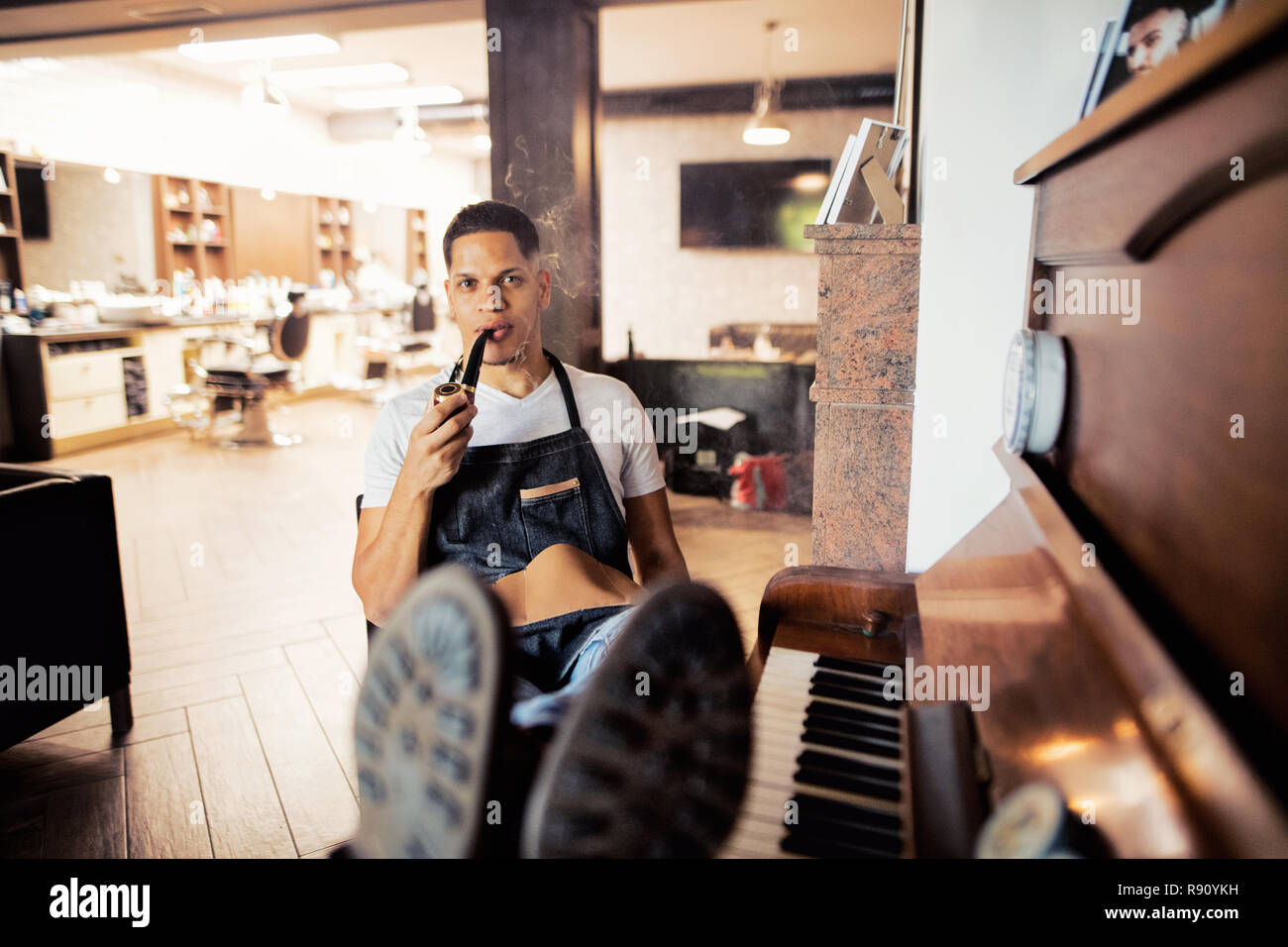 Young male hispanic haidresser and hairstylist sitting in barber shop, smoking a pipe and holding a drink. Stock Photo