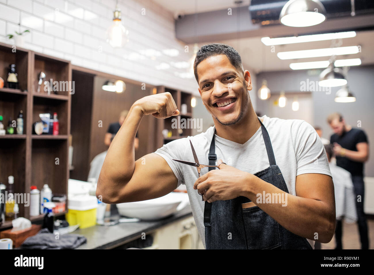 Young handsome hispanic haidresser and hairstylist standing in barber shop, flexing muscles. Stock Photo
