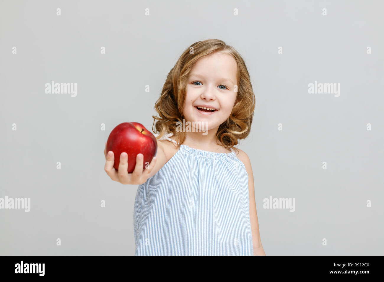 Portrait of a happy smiling little blonde girl on a gray background. A child holds out a red apple Stock Photo