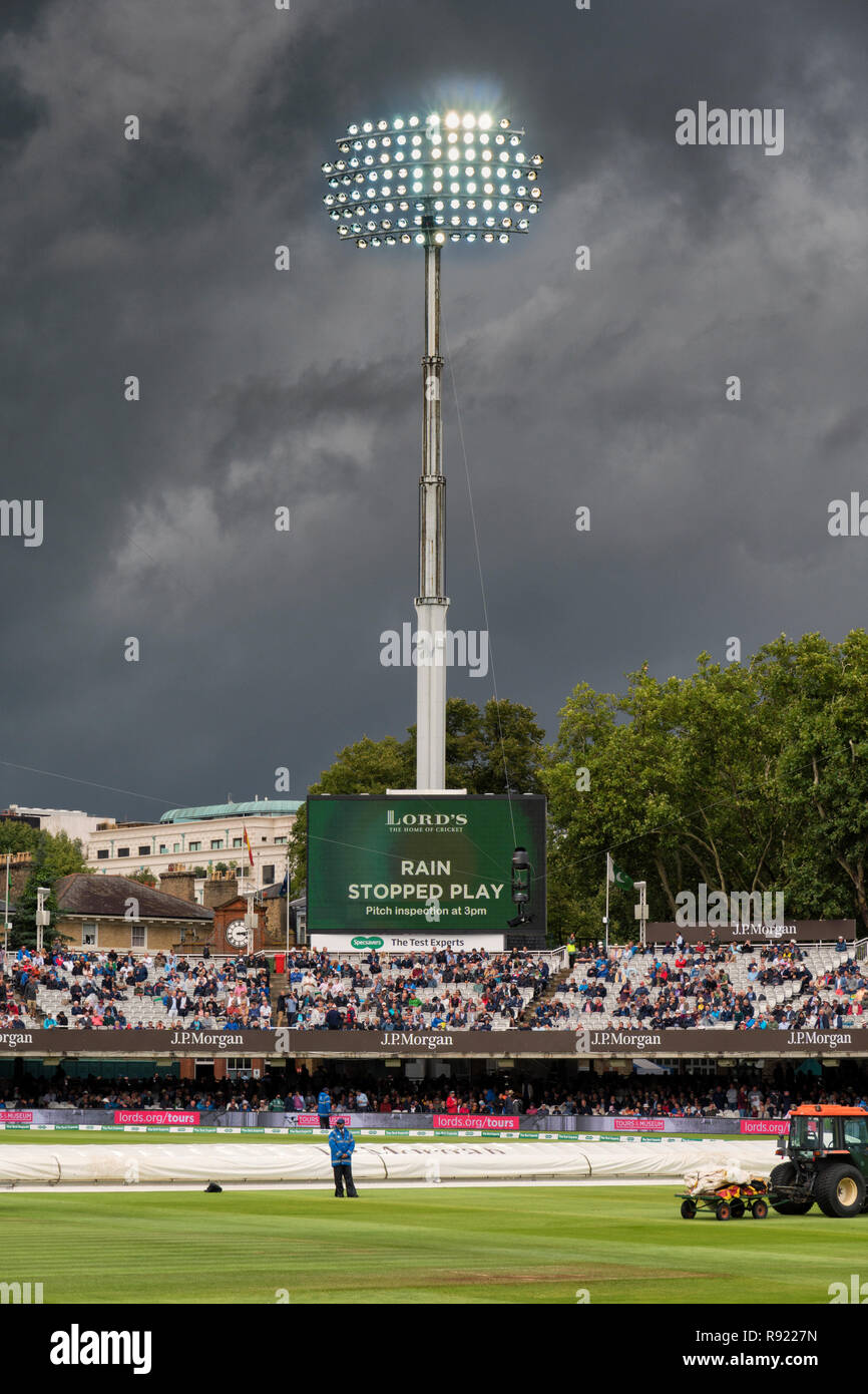 After heavy rain during a test match at Lords, the floodlights blaze brightly against an angry grey sky. A steward guards the covered pitch Stock Photo