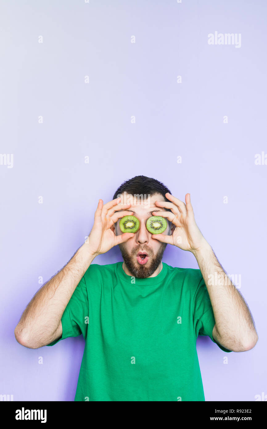 Young bearded holding slices of green kiwi fruit in front of his eyes, surprised and shocked. Light purple background, vertical orientation, copy spac Stock Photo
