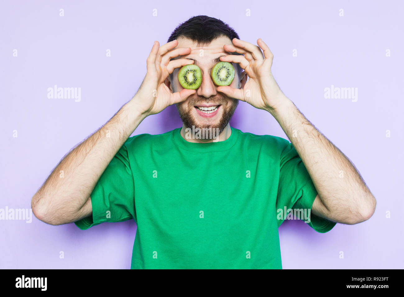 Young bearded man holding slices of green kiwi fruit in front of his eyes, smiling and surprised. Light purple background, copy space. Stock Photo
