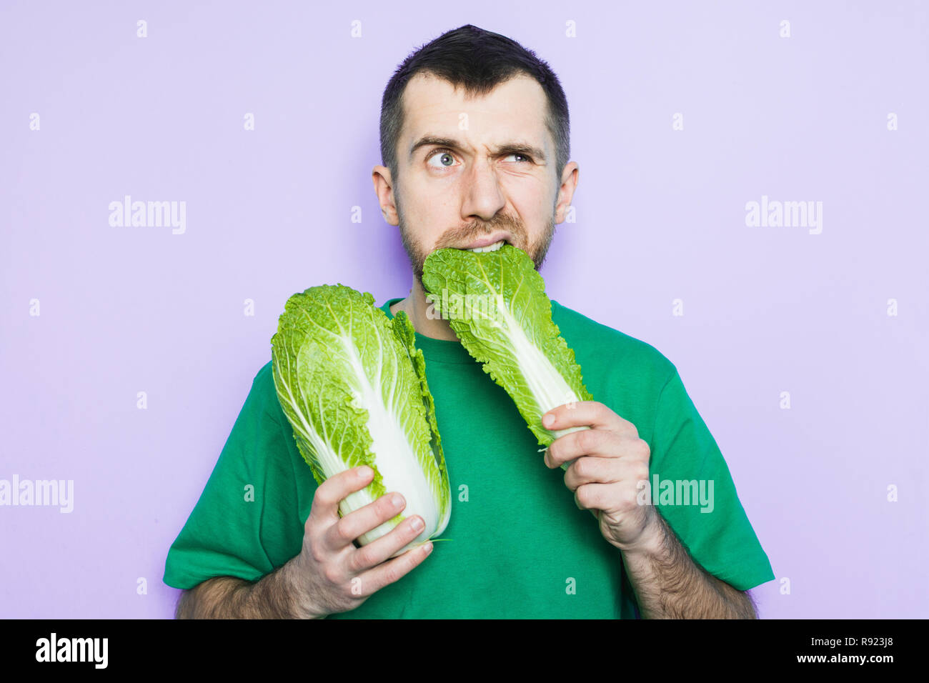 Young man biting on a leaf of Beijing napa cabbage, doubt face expression. Light purple background. Stock Photo