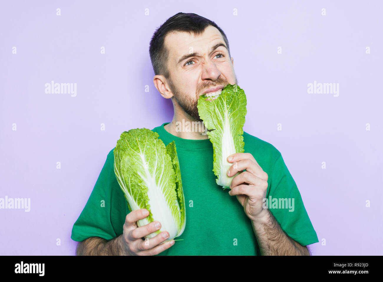 Young man biting on a leaf of Beijing napa cabbage, doubt face expression. Light purple background. Stock Photo
