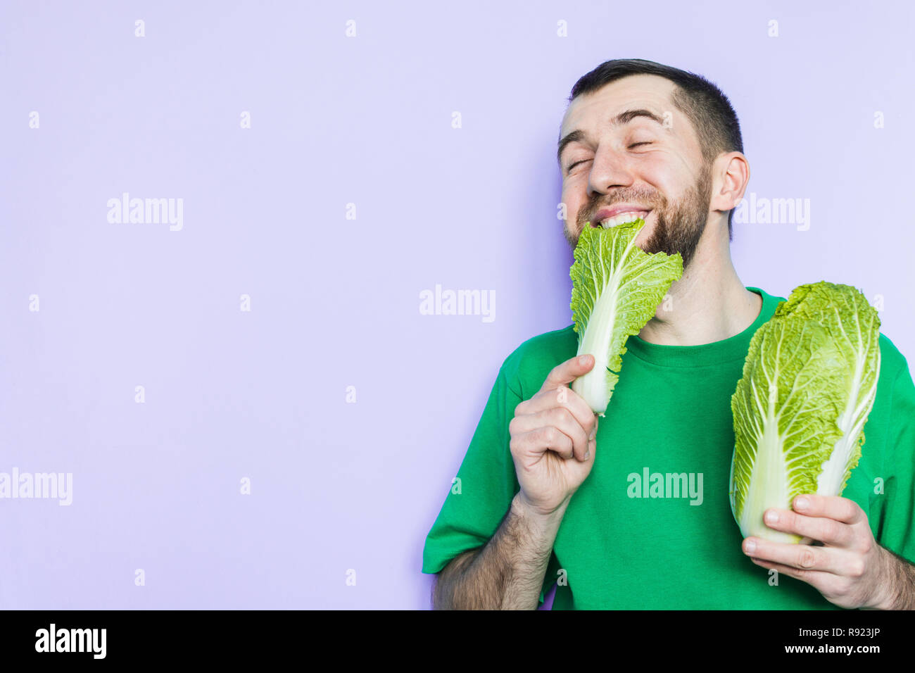 Young man biting on a leaf of Beijing napa cabbage, enjoyment face expression. Light purple background, copy space. Stock Photo