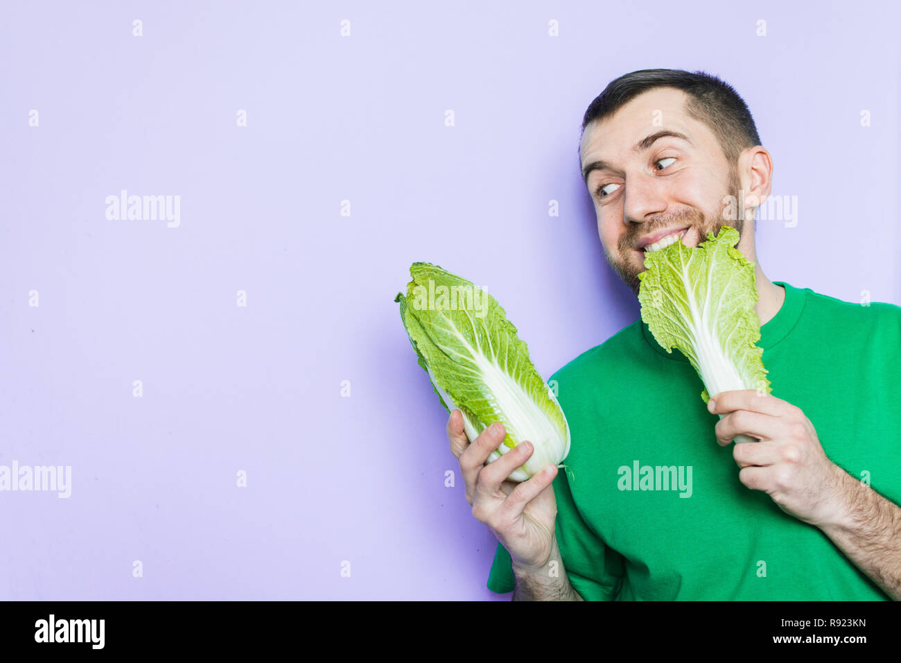Young man biting on a leaf of Beijing napa cabbage, enjoyment face expression. Light purple background, copy space. Stock Photo