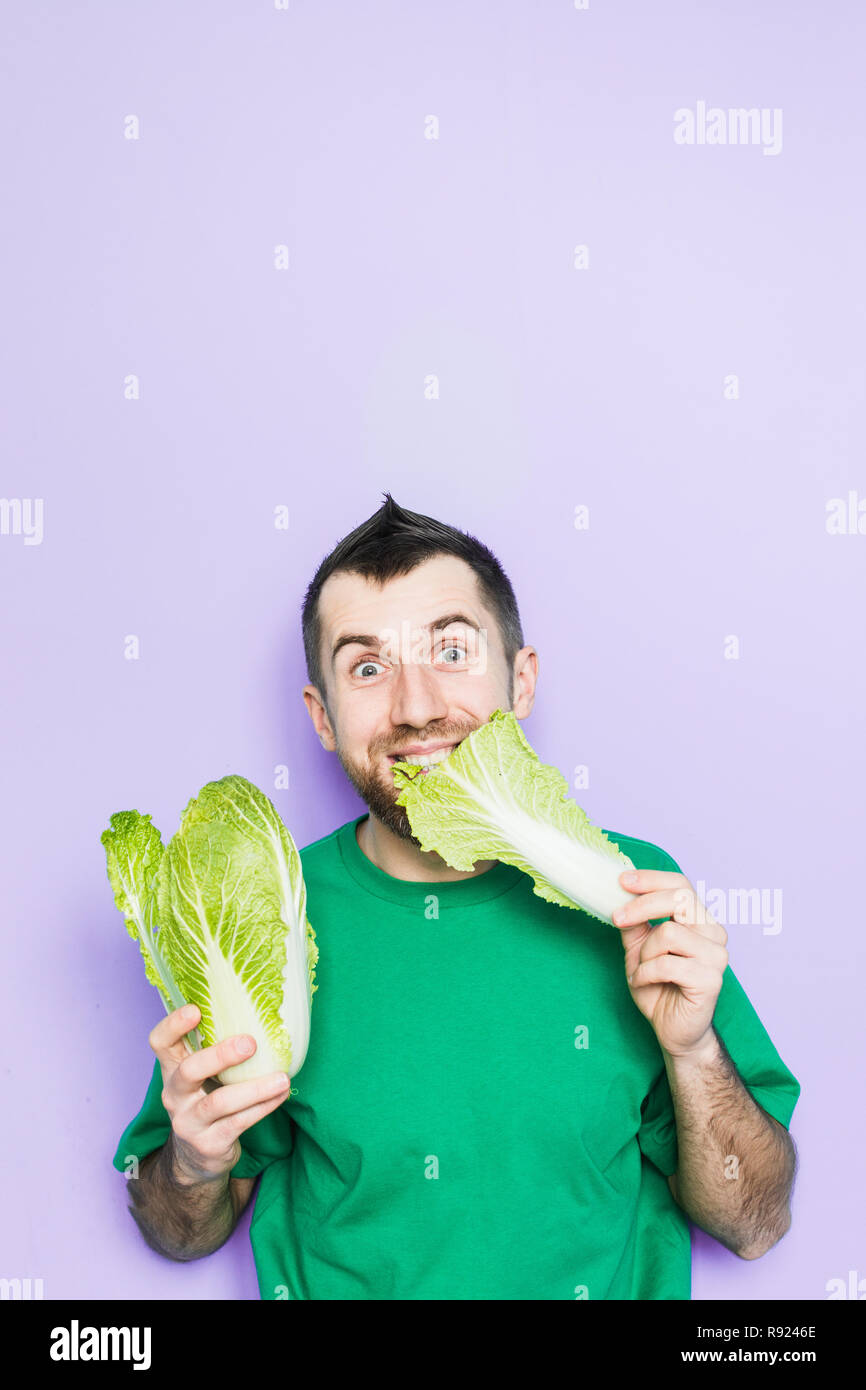 Young man biting on a leaf of Beijing napa cabbage, enjoyment face expression. Light purple background, copy space, vertical orientation. Stock Photo