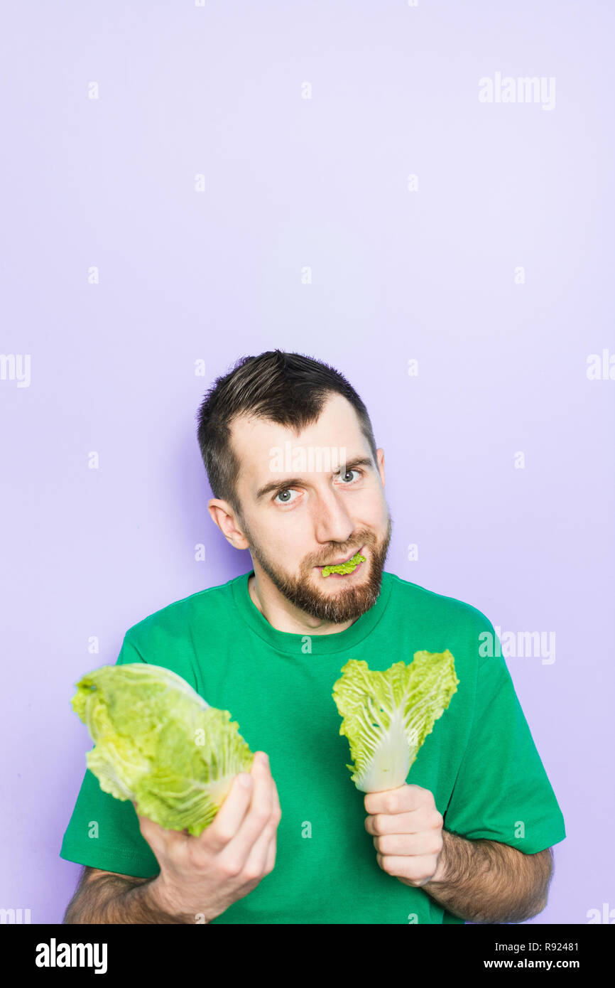 Young man biting on a leaf of Beijing napa cabbage, offering the product to the viewer. Light purple background, copy space, vertical orientation. Stock Photo