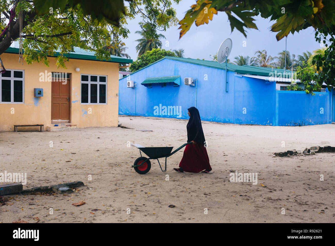 Woman wearing traditional hijab pushing wheelbarrow on sand in front of houses, Male, Thulusdhoo, Maldives Stock Photo