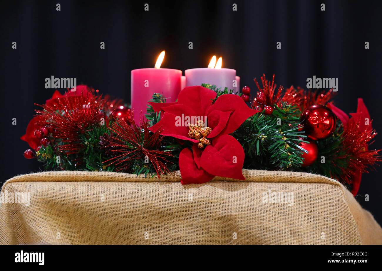 A red and green imitation Christmas wreath decoration with tinsel and poinsettia flowers encircling large lit candles and flame. Xmas concept Stock Photo