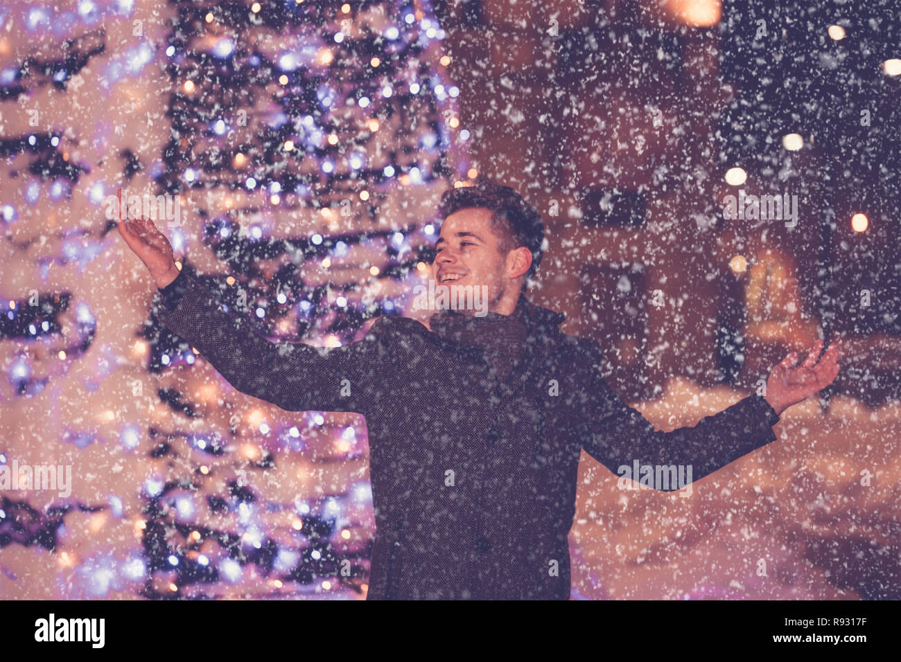 Free young man with outstretched arms looking up while being excited about winter weather at decorated city street at night, he standing under snow -  Stock Photo