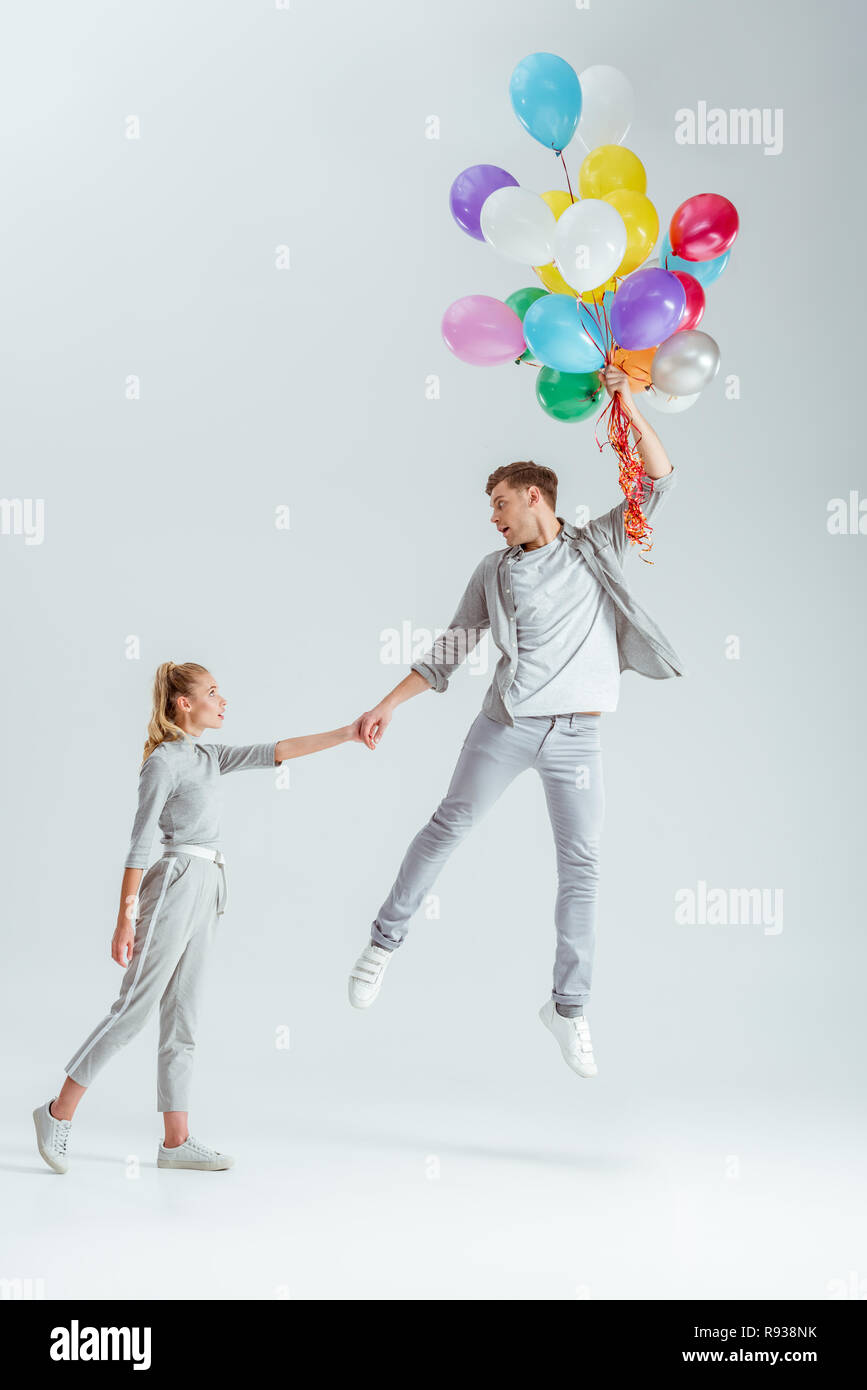 beautiful woman holding hand of man jumping in air with bundle of colorful balloons on grey background Stock Photo