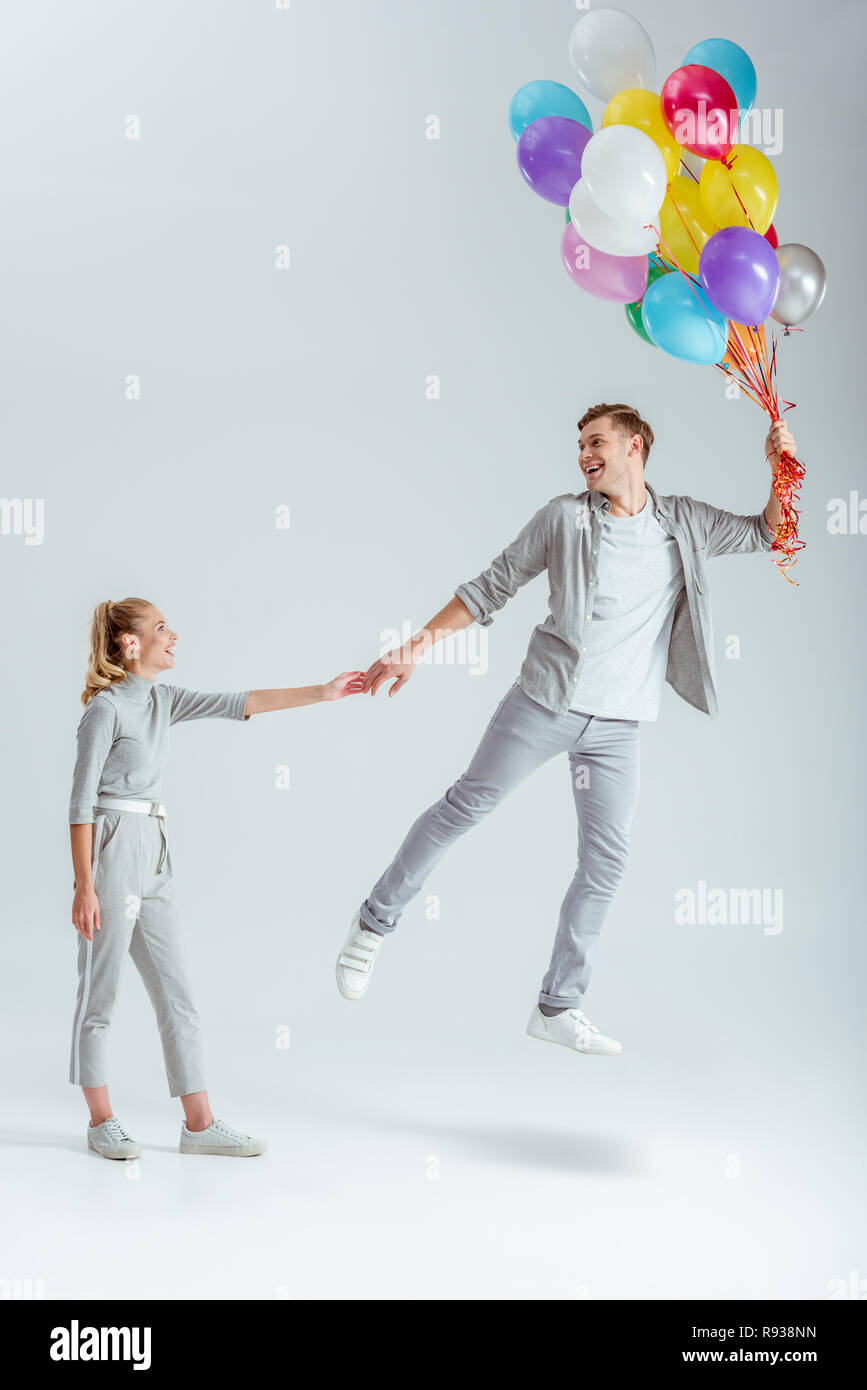 woman in grey clothing holding hand of happy man jumping in air with bundle of colorful balloons on grey background Stock Photo