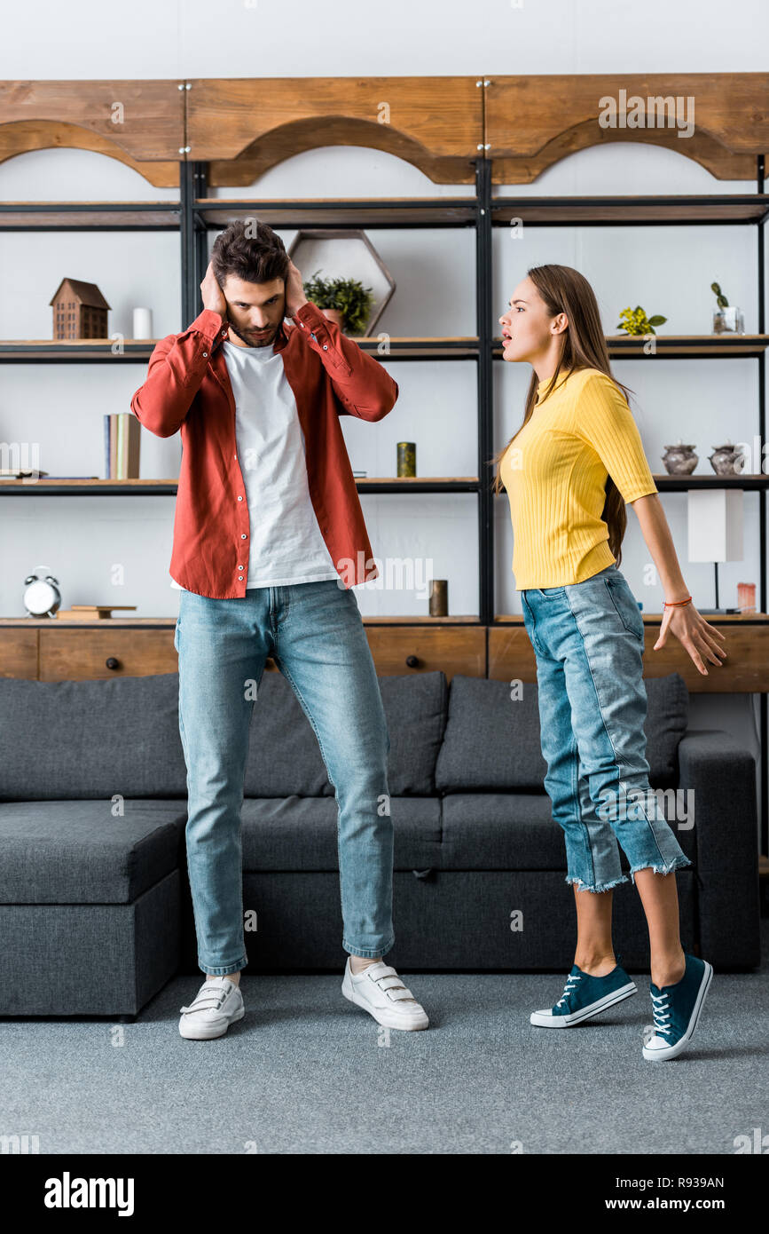 angry girlfriend screaming at boyfriend while man putting hands on ears in living room Stock Photo