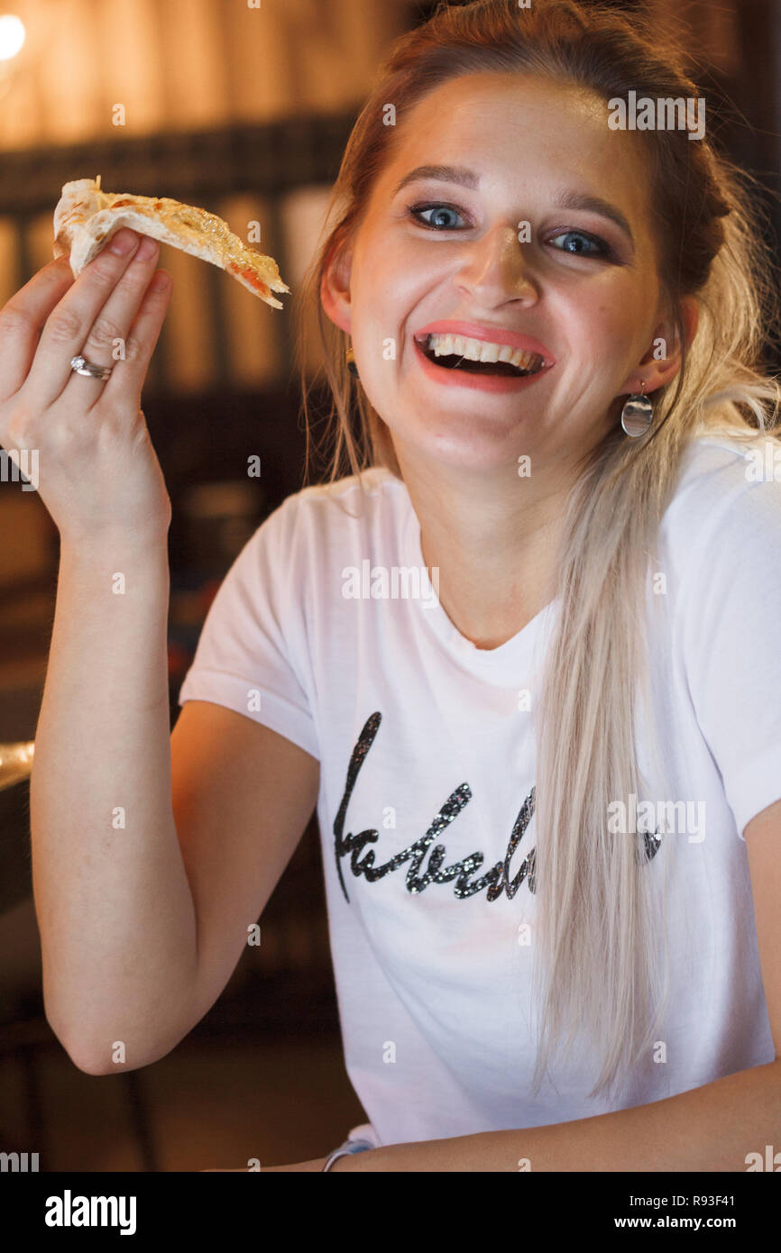 Casual girl eating pizza in the restaurant and enjoying this. Funny blonde girl in white t-shirt eating pizza at restaurant Stock Photo