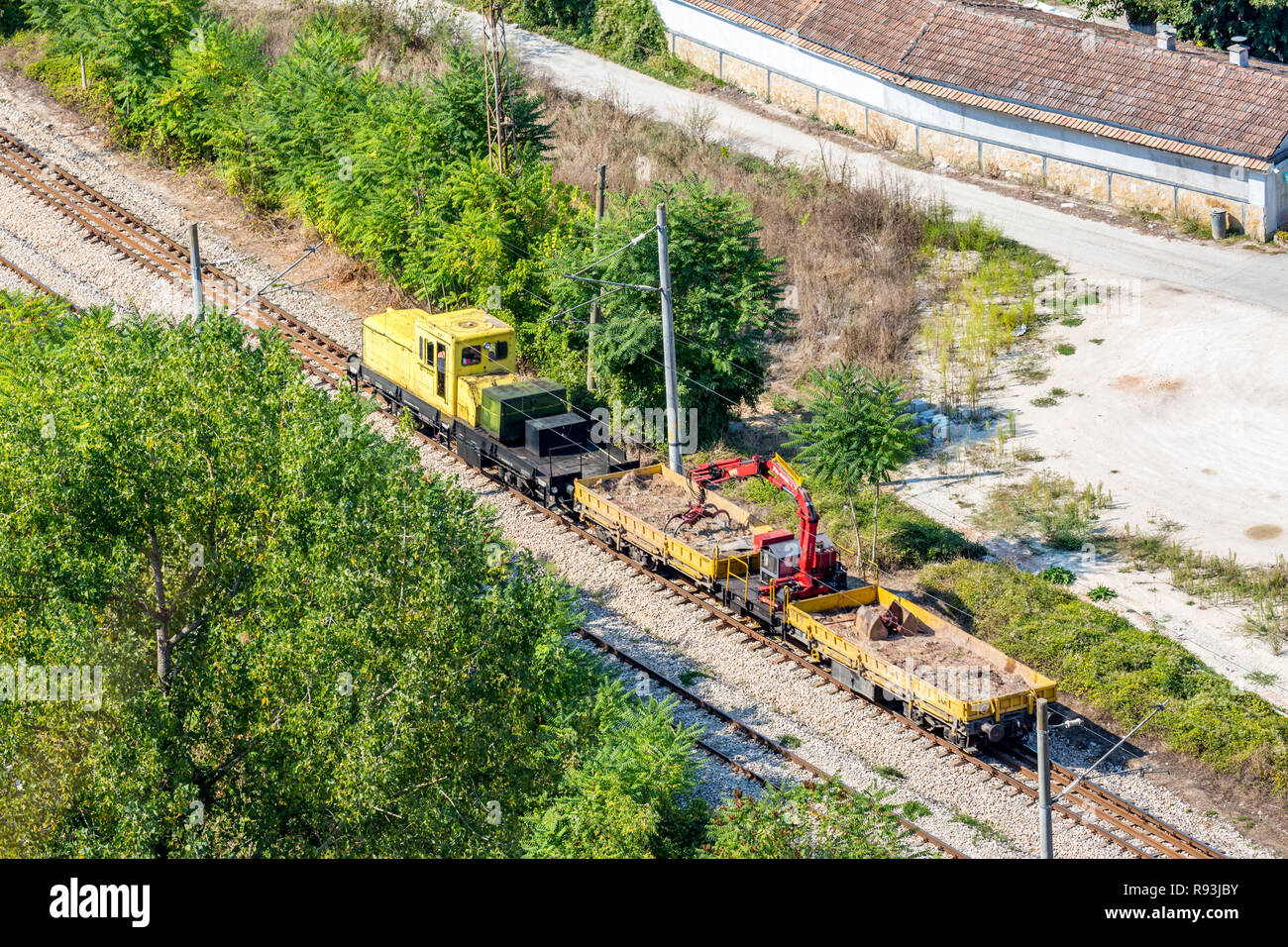 Yellow railway service diesel engine moving on rail line dragging two wagons with pebbles and small red crane going to construction site. Rural Bulgar Stock Photo