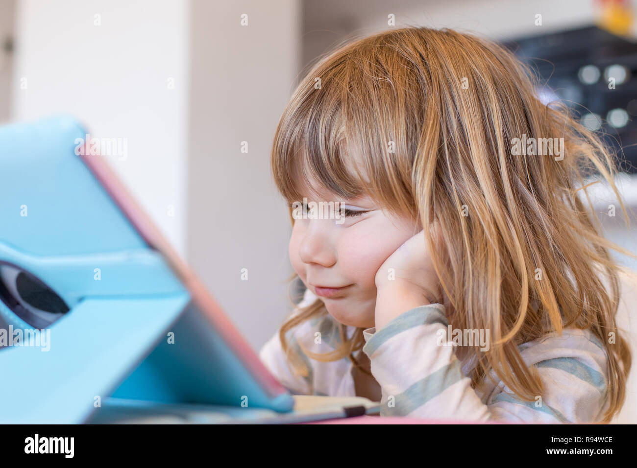 funny face expression of three years old blonde child sitting indoor, with head over hand watching digital tablet on table Stock Photo