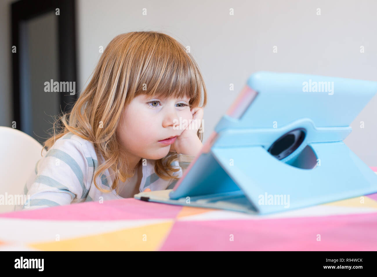 funny face expression of three years old blonde child sitting indoor, with head over hand watching digital tablet on table Stock Photo