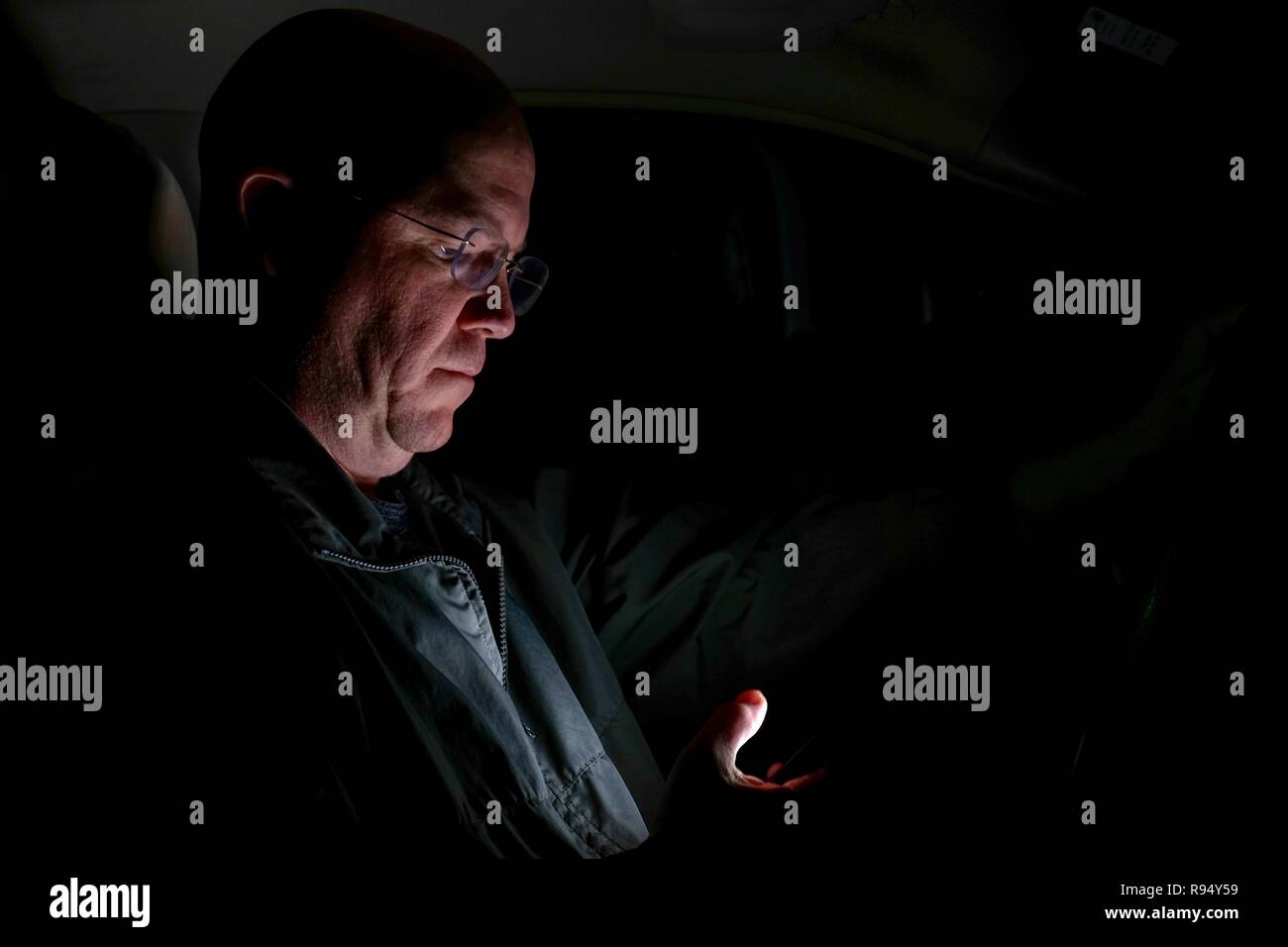 A caucasion man pretends to engage in the reckless act of texting while driving a vehicle, with the glow of the screen on his face in the dark. Stock Photo