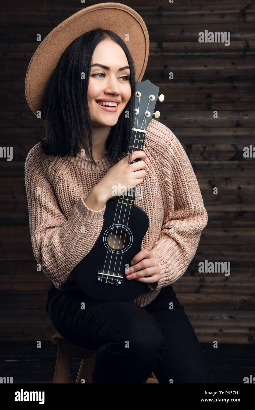 Young smiling girl with a ukulele sit in studio on a wooden background. Stock Photo