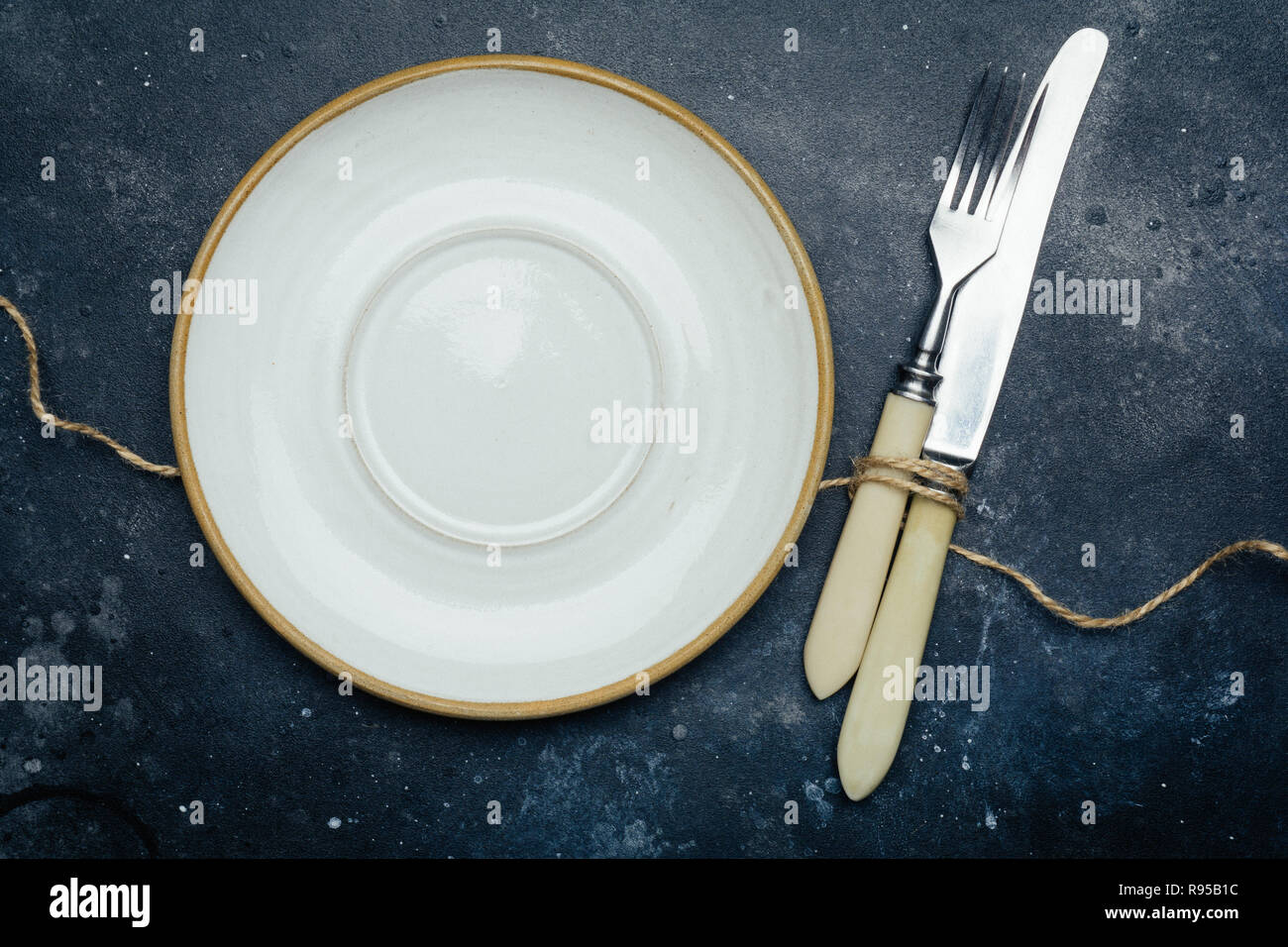 Empty white plate (ceramic, rustic) on a dark blue background with a knife and fork. Copy space. Stock Photo