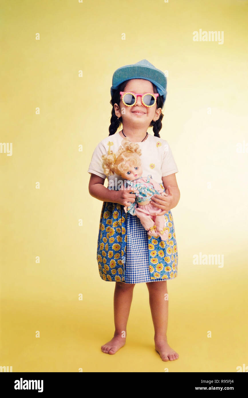 Girl wearing cap, fancy goggles, doll in hand, standing on yellow