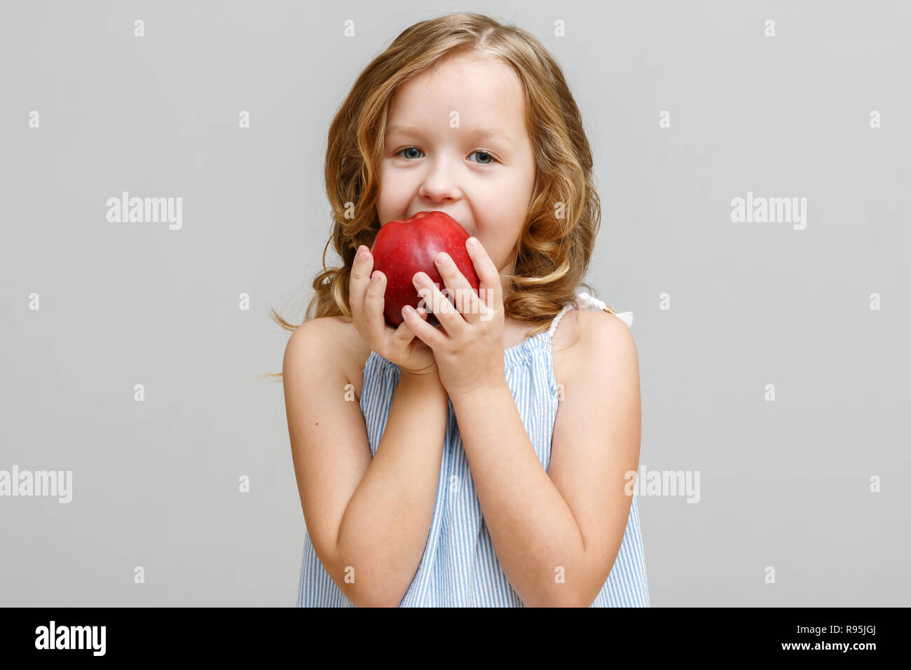 Portrait of a happy smiling little blonde girl on a gray background. Baby bites red apple Stock Photo