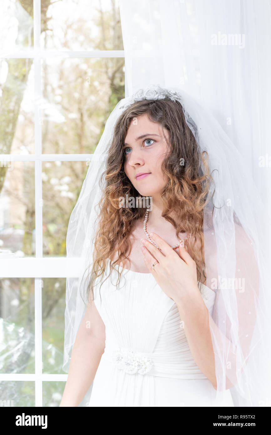 Closeup portrait of young female person, woman, bride in wedding dress, veil, face, pearl necklace, hair, standing, looking up by glass window, white  Stock Photo