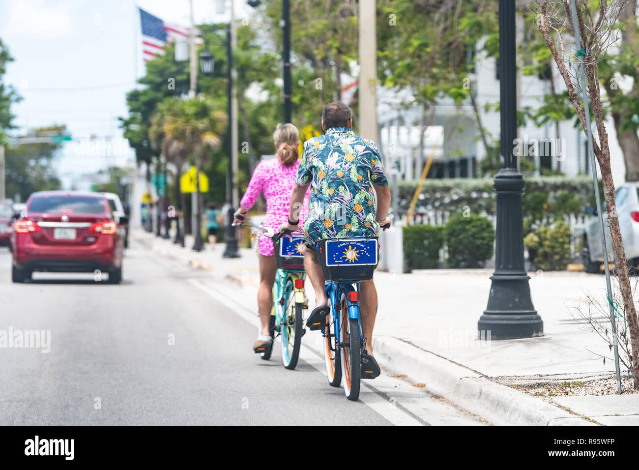 Key West, USA - May 1, 2018: Back of couple, man, woman riding rental bikes, bicycles with conch republic flag on Truman Avenue street, road, cars in  Stock Photo