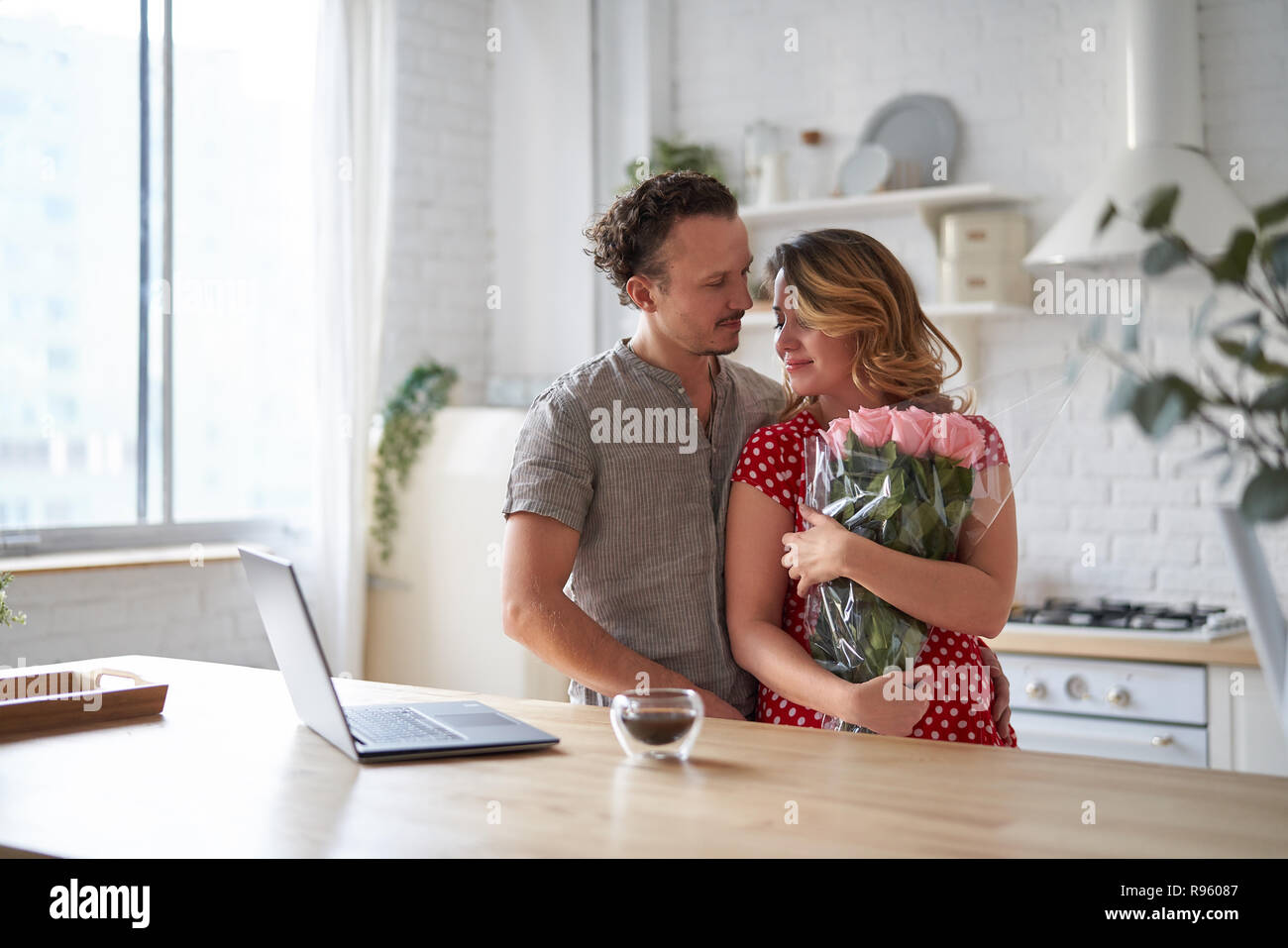Surprise. Beautiful romantic couple in kitchen. Young man is presenting flowers to his beloved. Feel of happiness. Stock Photo