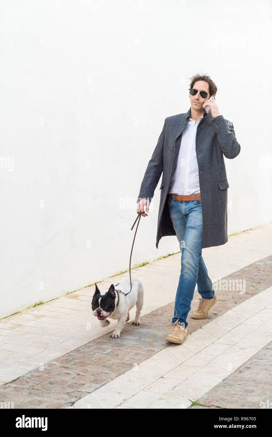 Urban scene. Man dressed in casual clothes walking his dog down the street while talking on the smartphone. Stock Photo