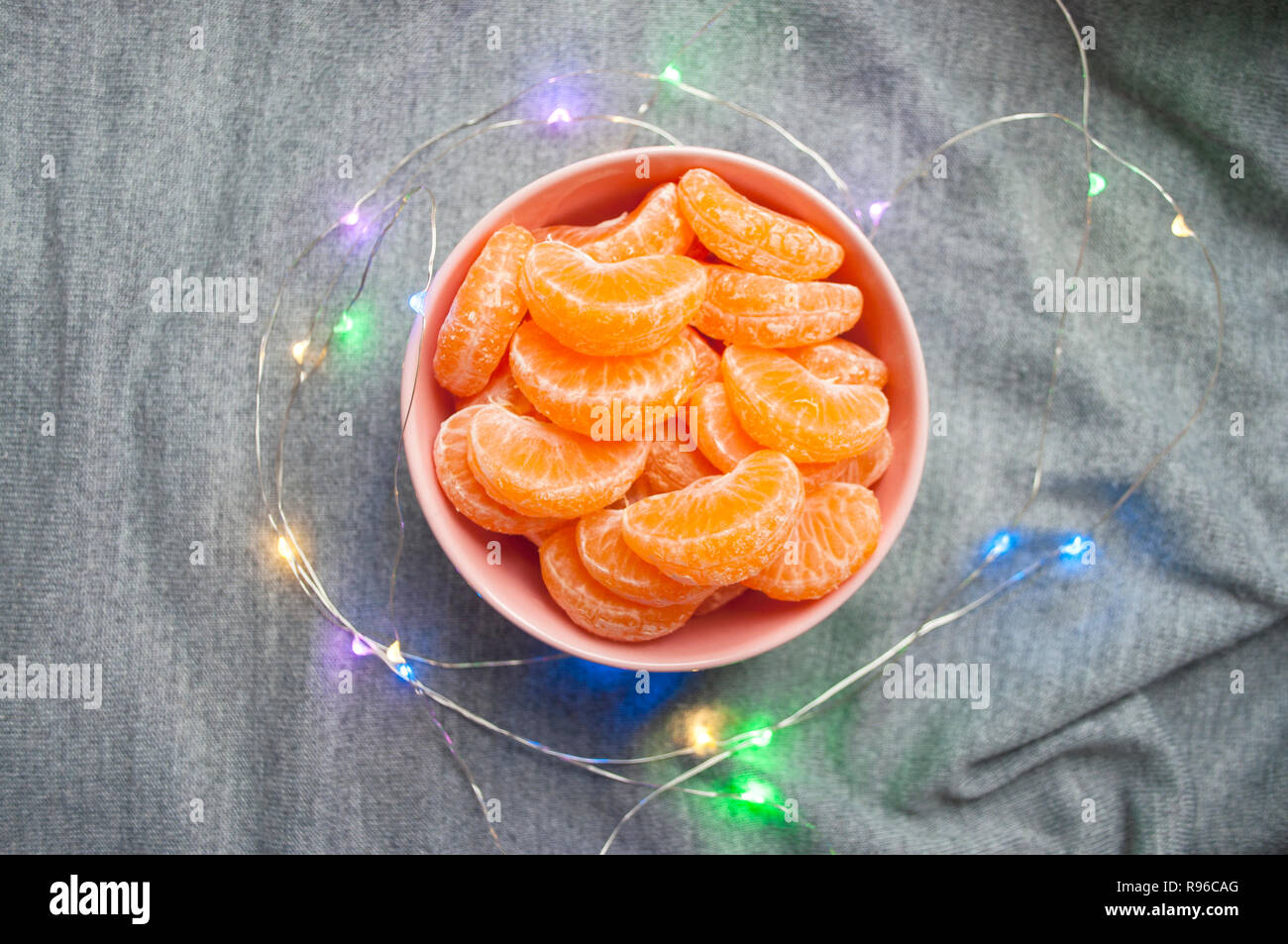 Flat lay of pink bowl full of peeled sweet tangerines on grey background with garland lights. Stock Photo