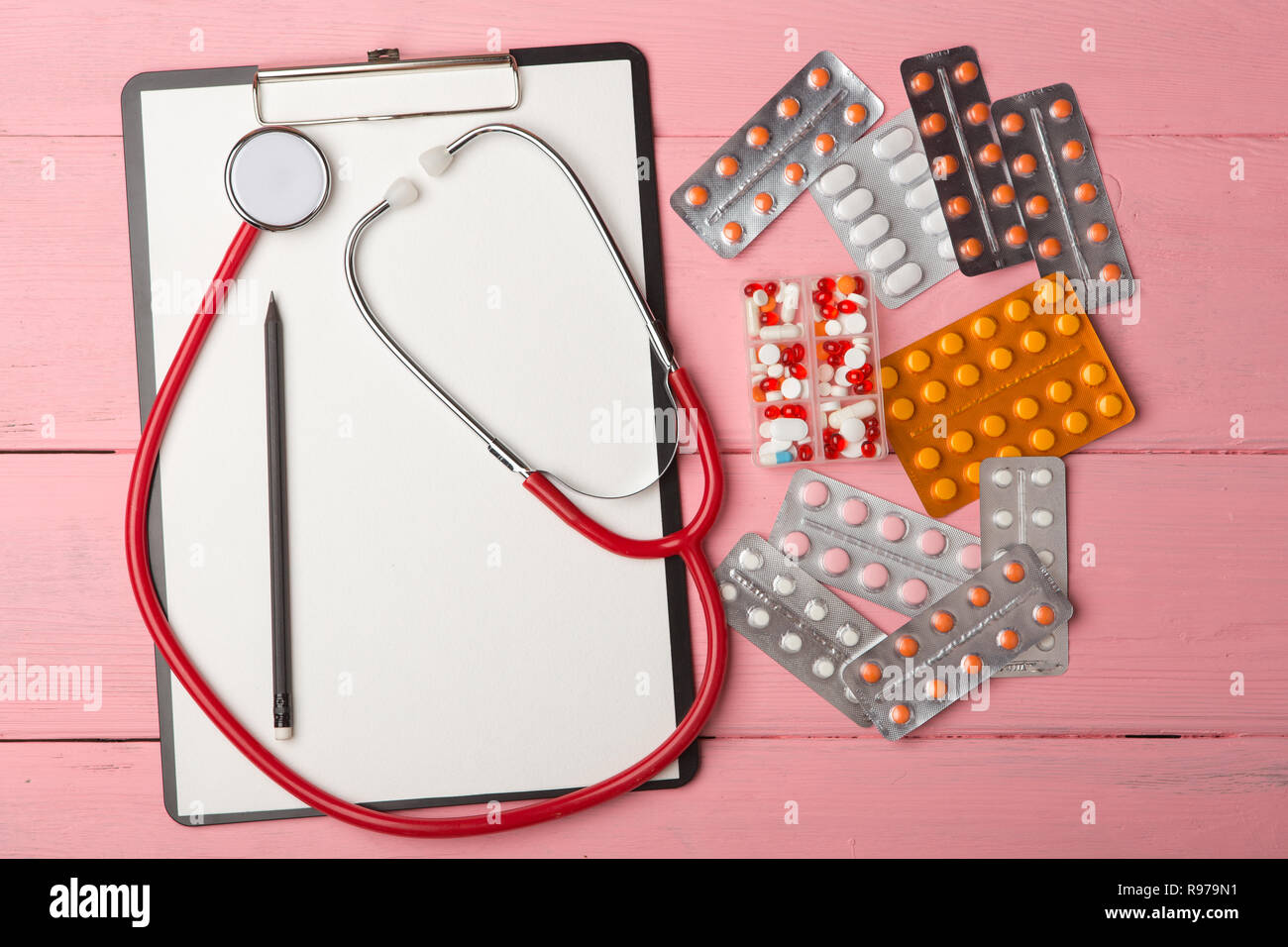 Doctor workplace with blank clipboard, medicine red stethoscope and pills on pink wooden table. Medication prescription. Health care or illness Stock Photo