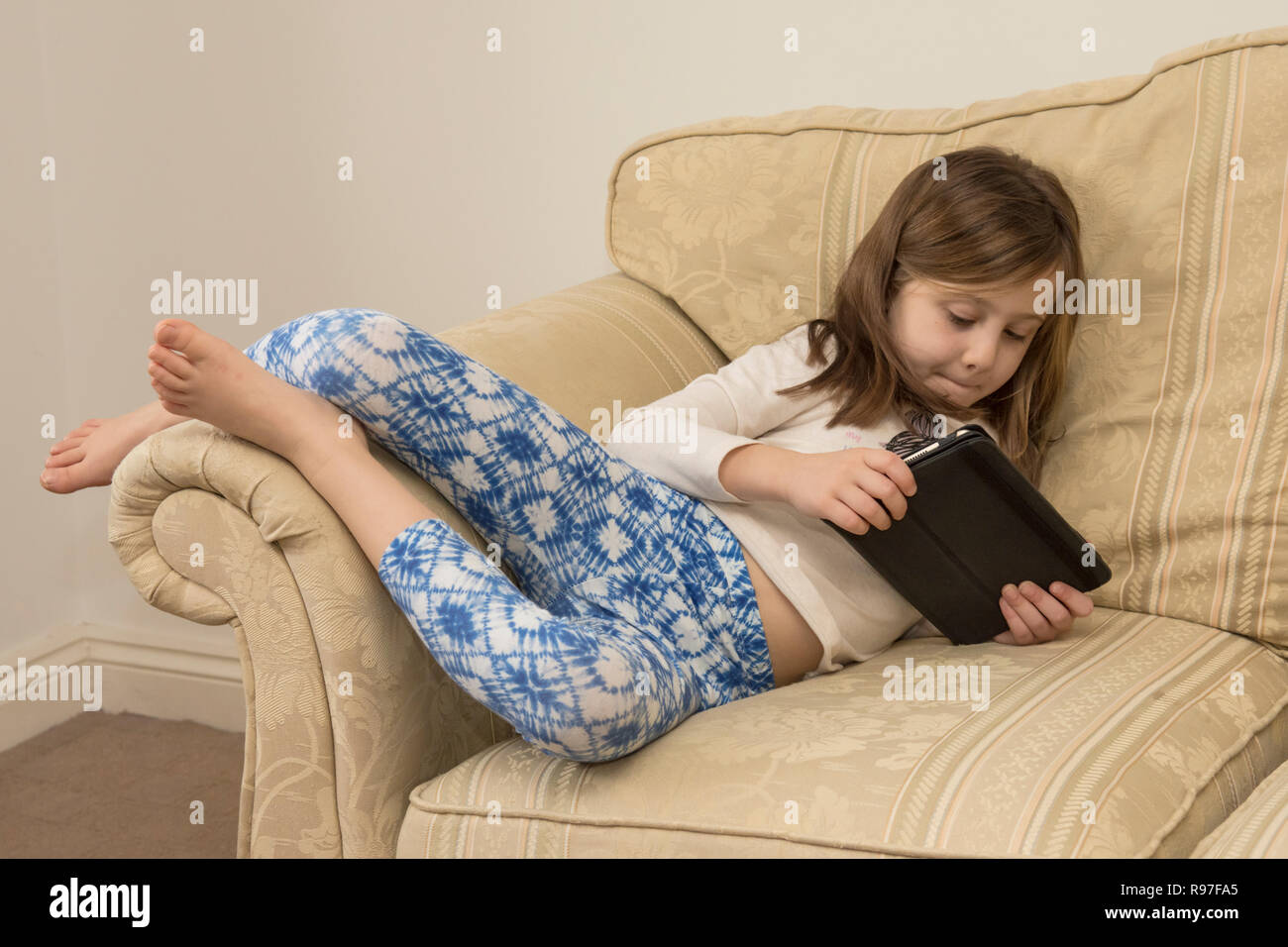 young girl laying of a sofa playing with an iPad, digital device, tablet, electronic media, social media, six-year-old. Stock Photo
