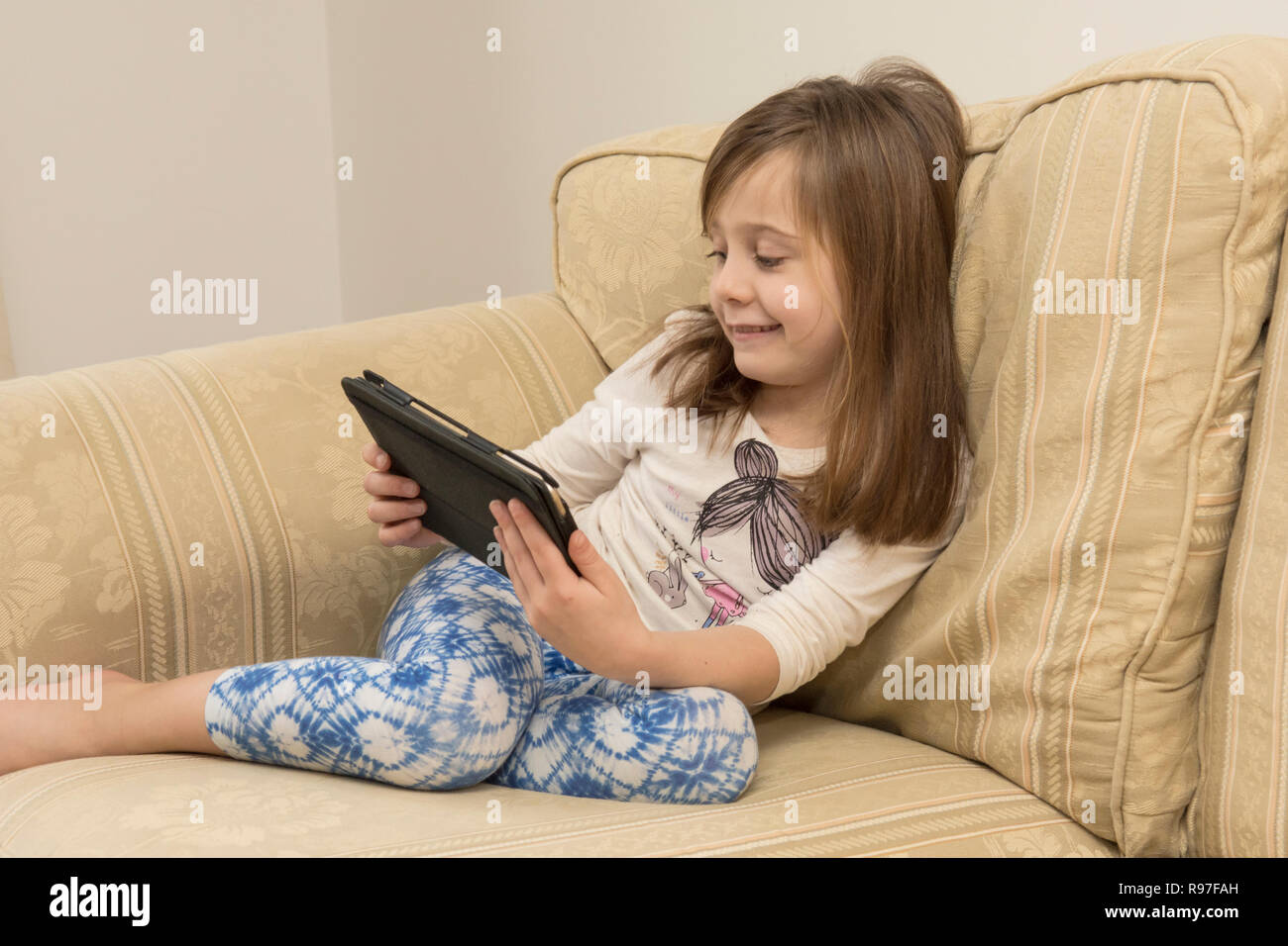 young girl laying of a sofa playing with an iPad, digital device, tablet, electronic media, social media, six-year-old. Stock Photo