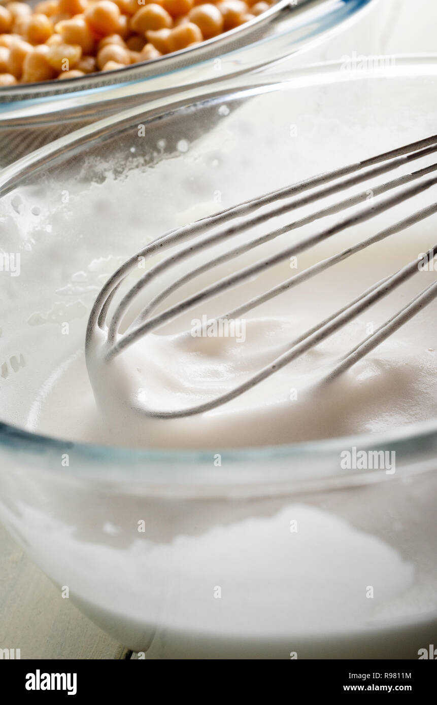 Glass bowl of whisked aquafaba (chickpea water) in foreground, with the drained pulses in background. Stock Photo