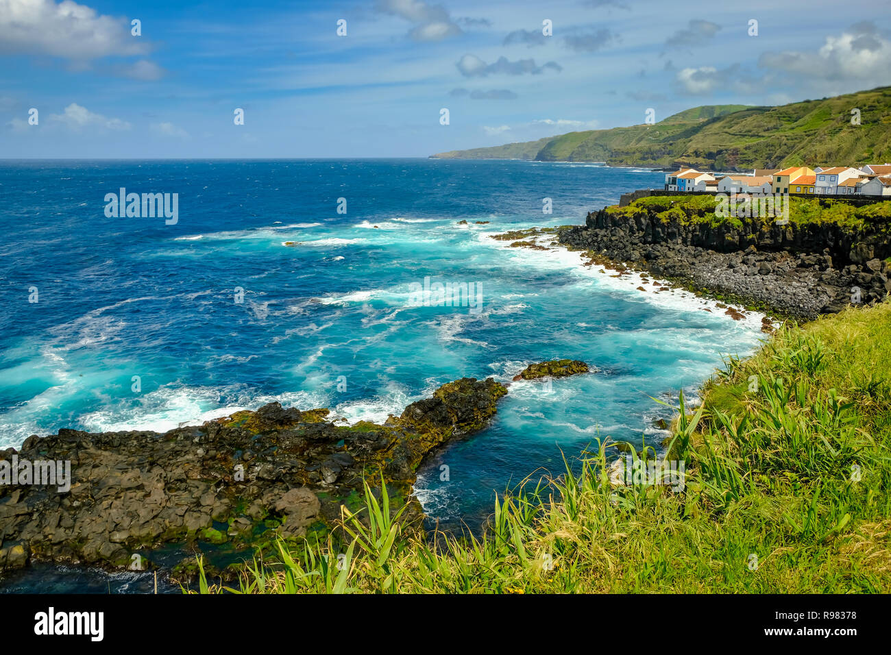 Rocky black coast of the Azores with crashing blue waves and a tiny little village on a beautiful sunny day. Stock Photo