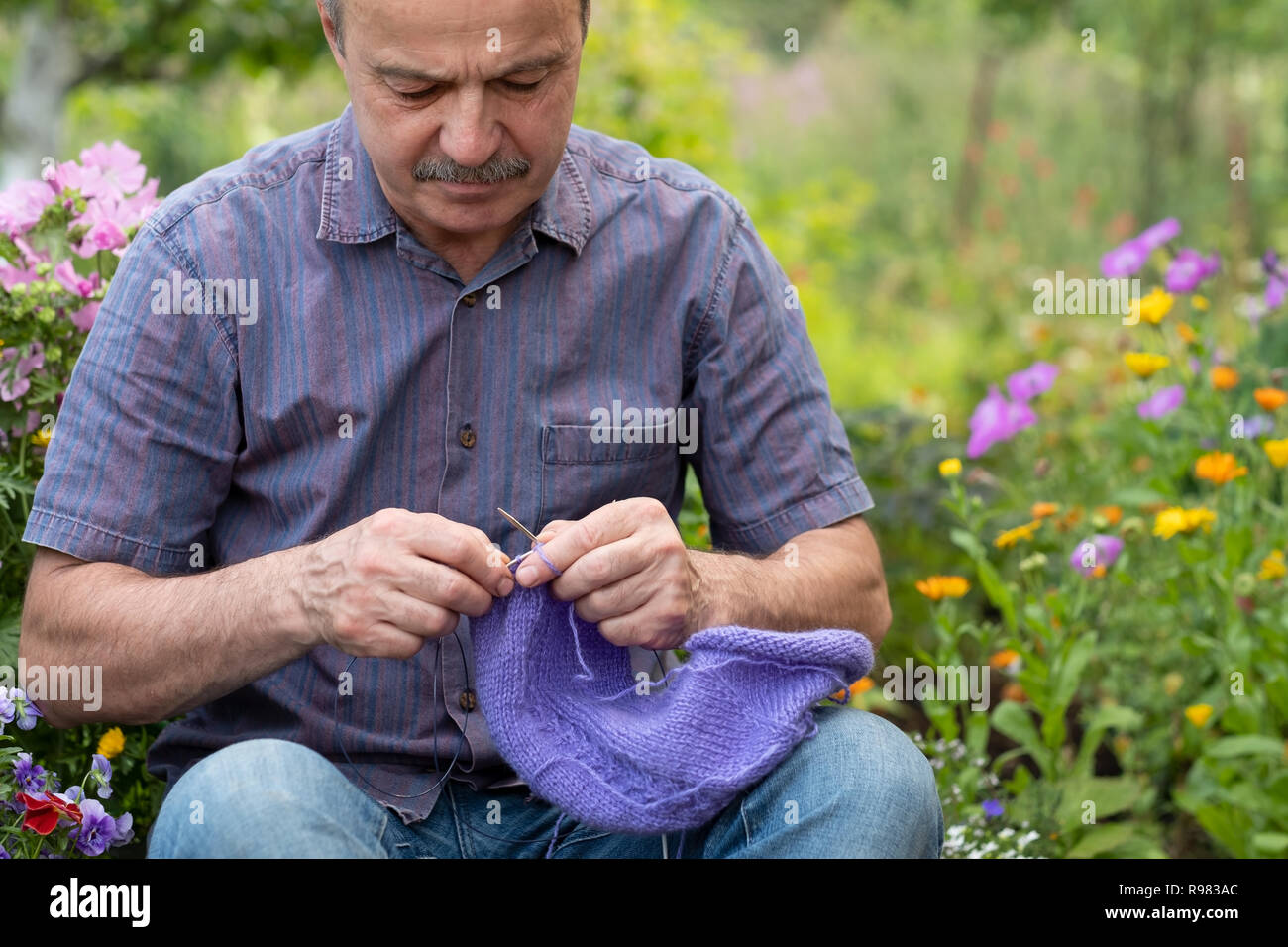 Old handsome man sitting at his summer garden with the knitting needles. Stock Photo